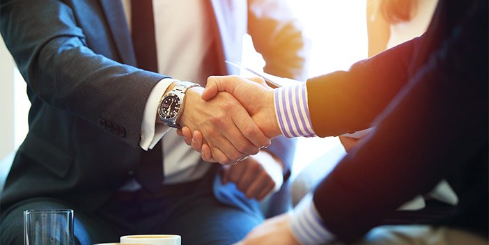 Two people dressed in suits, shaking hands across a table.
