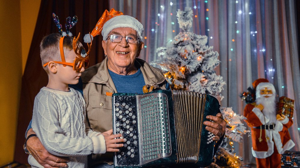 An older man smiling wearing a Santa hat and playing an accordion with a young boy who is wearing reindeer shaped glasses. They are both sat in front of a Christmas tree with warm twinkling lights.