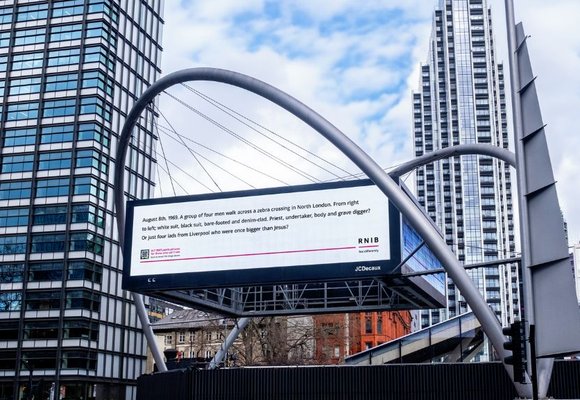 A digital billboard in between two glass skyscrapers in London. Black text on a white background on the billboard reads 'August 8th, 1969. A group of four men walk across a zebra crossing in North London. From right to left, white suit, black suit, bare-footed and denim-clad. Priest, undertaker, body and grave differ? Or just four lads from Liverpool who were once bigger than Jesus?'